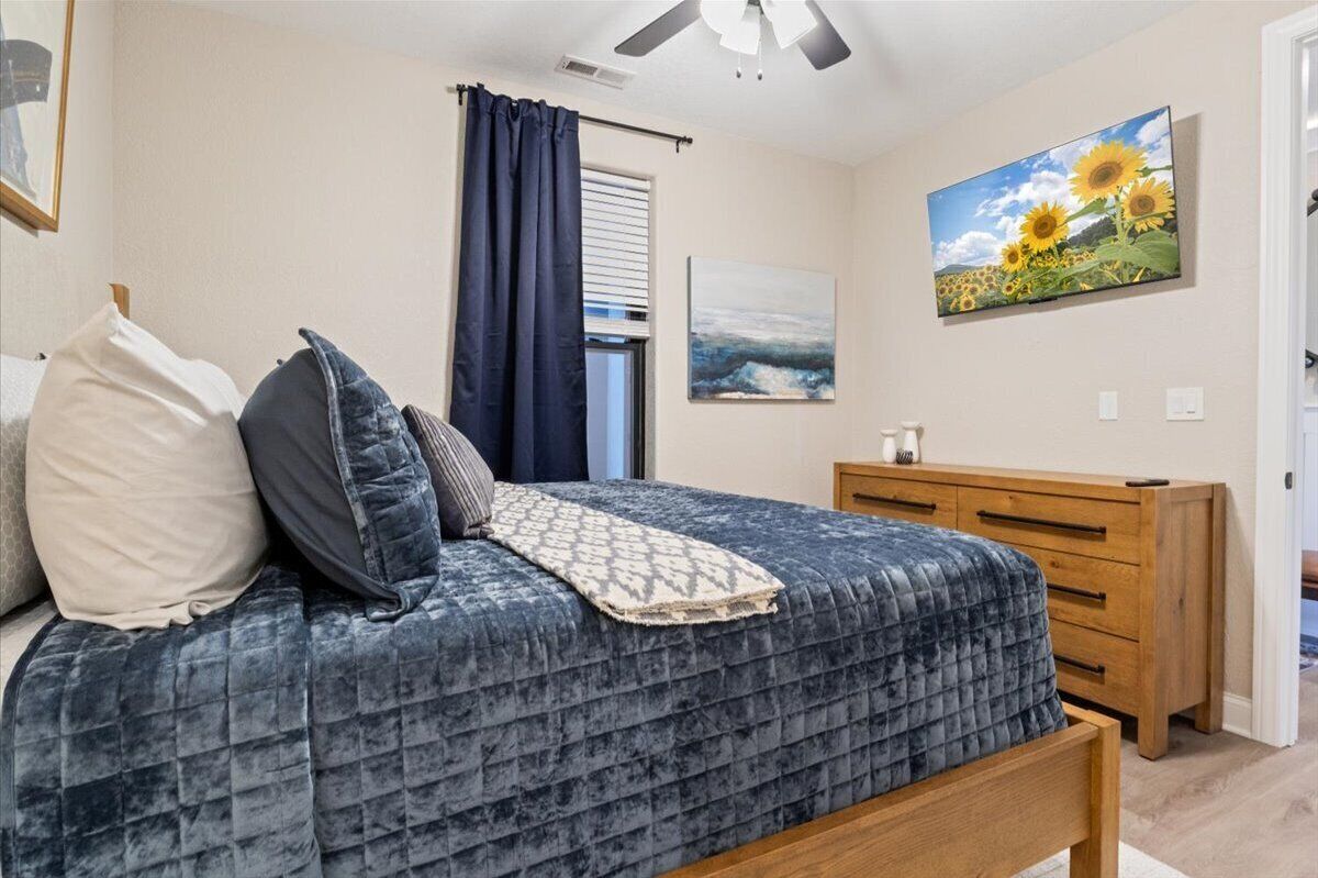 Une chambre à coucher avec un lit, une commode en bois, un ventilateur de plafond, une fenêtre avec des rideaux et un tableau représentant des tournesols.