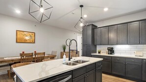 A modern kitchen with dark cabinets, a white countertop, and a built-in sink. There is a dining area with wooden chairs and a table, and a pendant light above the kitchen counter. - Family-fun Lakeside Luxury at Topsider in Osage w/ 2 Pools, Prime Views Pack N' Play & Games (Osage Beach)