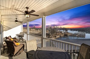 A covered balcony with wicker furniture, a table, and a view of a river and construction site during sunset. - Beautiful Cove Views & Plenty Relaxing Spaces (Sunrise Beach)