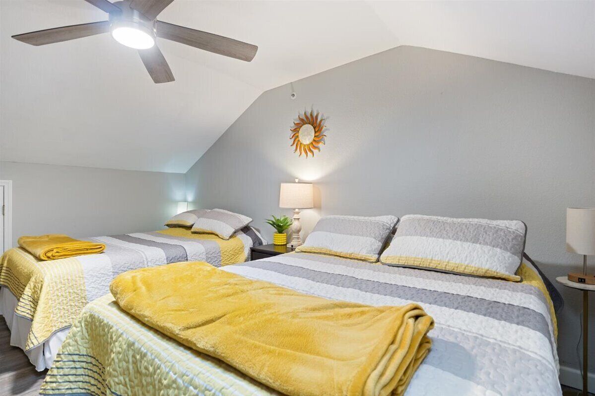 A bedroom with two beds, a ceiling fan, a wall-mounted sun decoration, and a small table with a lamp.