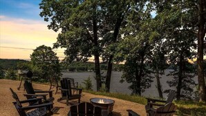 A lakeside seating area with Adirondack chairs and a fire pit, surrounded by trees and a view of the lake. - 17 Lakefront Acres, Private Dock, Firepit, Pool Table, Kayaks, Fishing (Gravois Mills)