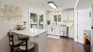 A modern kitchen with white cabinets, a central island, wooden chairs, and a view of the outdoors. - Minutes From the Strip - Nightlife and Dining! (Lake Ozark)