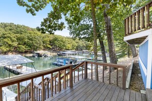 Une terrasse en bois surplombant une marina où sont amarrés des bateaux, le tout entouré d’arbres et de collines.