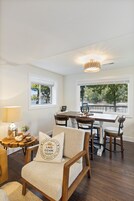 Una sala de estar acogedora con una silla de madera, una mesa de comedor y una araña. Se ven árboles y una valla a través de las ventanas.