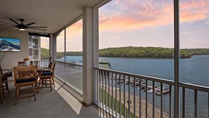Uma varanda com vista para um lago e uma floresta, uma área de jantar com cadeiras e uma mesa, e um ventilador de teto.