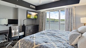 Quarto de hotel com cama, escrivaninha, cadeira, televisão e janela com cortinas.
