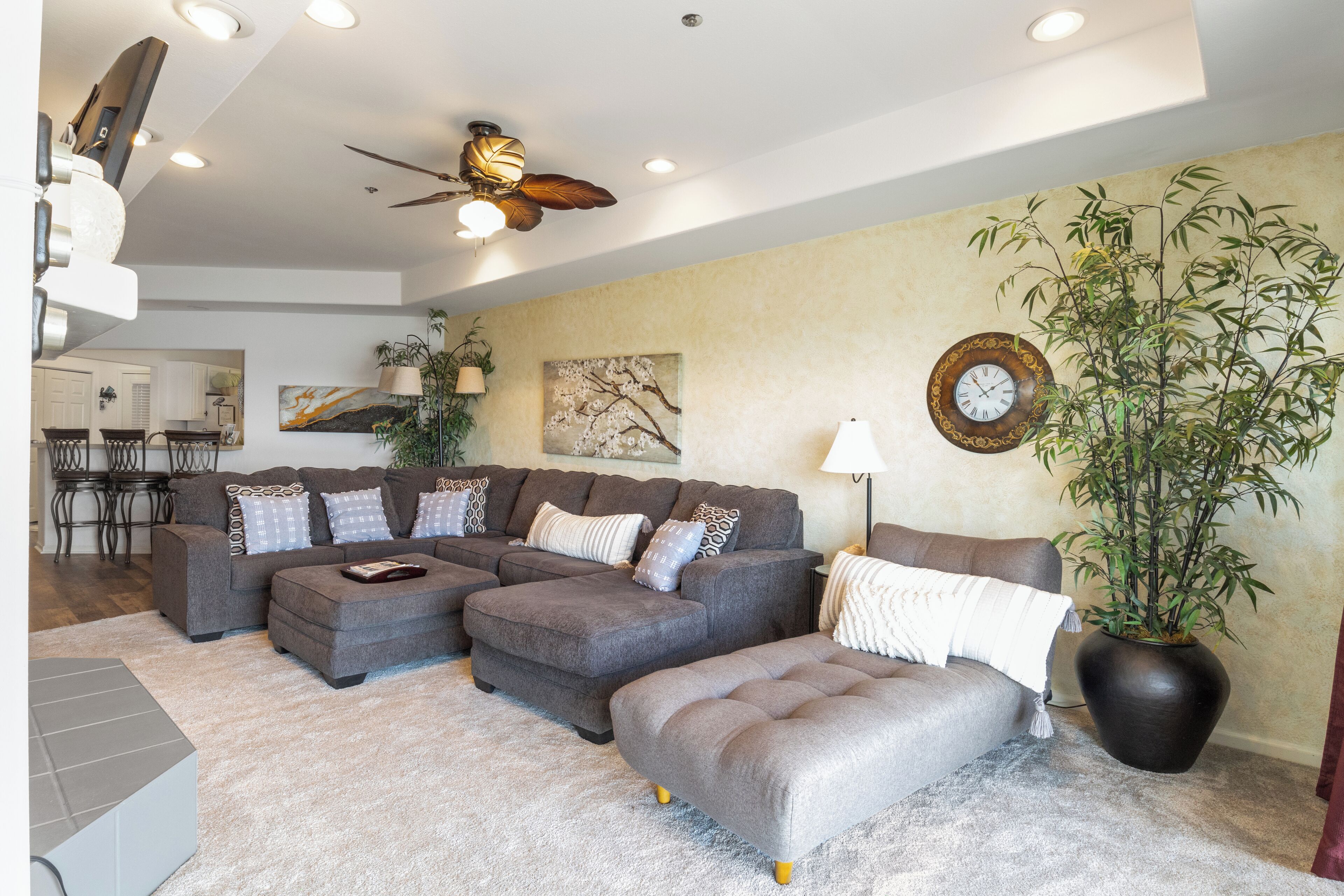 A modern living room with a sectional sofa, ottoman, ceiling fan, and a large potted plant.