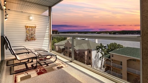 A balcony with a view of a river and buildings, a chair, and a decorative wall hanging. - Prime Spot, Breathtaking Views, Nearby Attractions (Lake Ozark)
