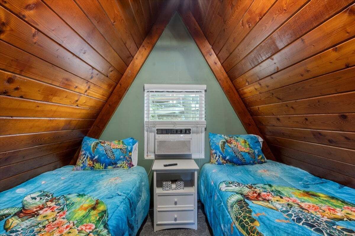 Ein schrägwandiges Schlafzimmer im Dachgeschoss mit Holzwänden. Es gibt zwei Betten mit passender Blumenbettwäsche, einen kleinen weißen Nachttisch und ein Fenster mit Jalousien.