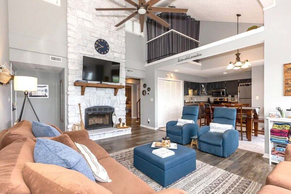 A spacious living room with a fireplace, a ceiling fan, and a dining area with a kitchen in the background. - 3 Bedroom Osage Beach Condo w/ Pool & Main Channel Views (Osage Beach)