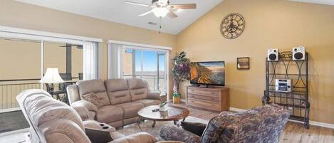 A spacious living room with a ceiling fan, a large window, a television, and a speaker system.