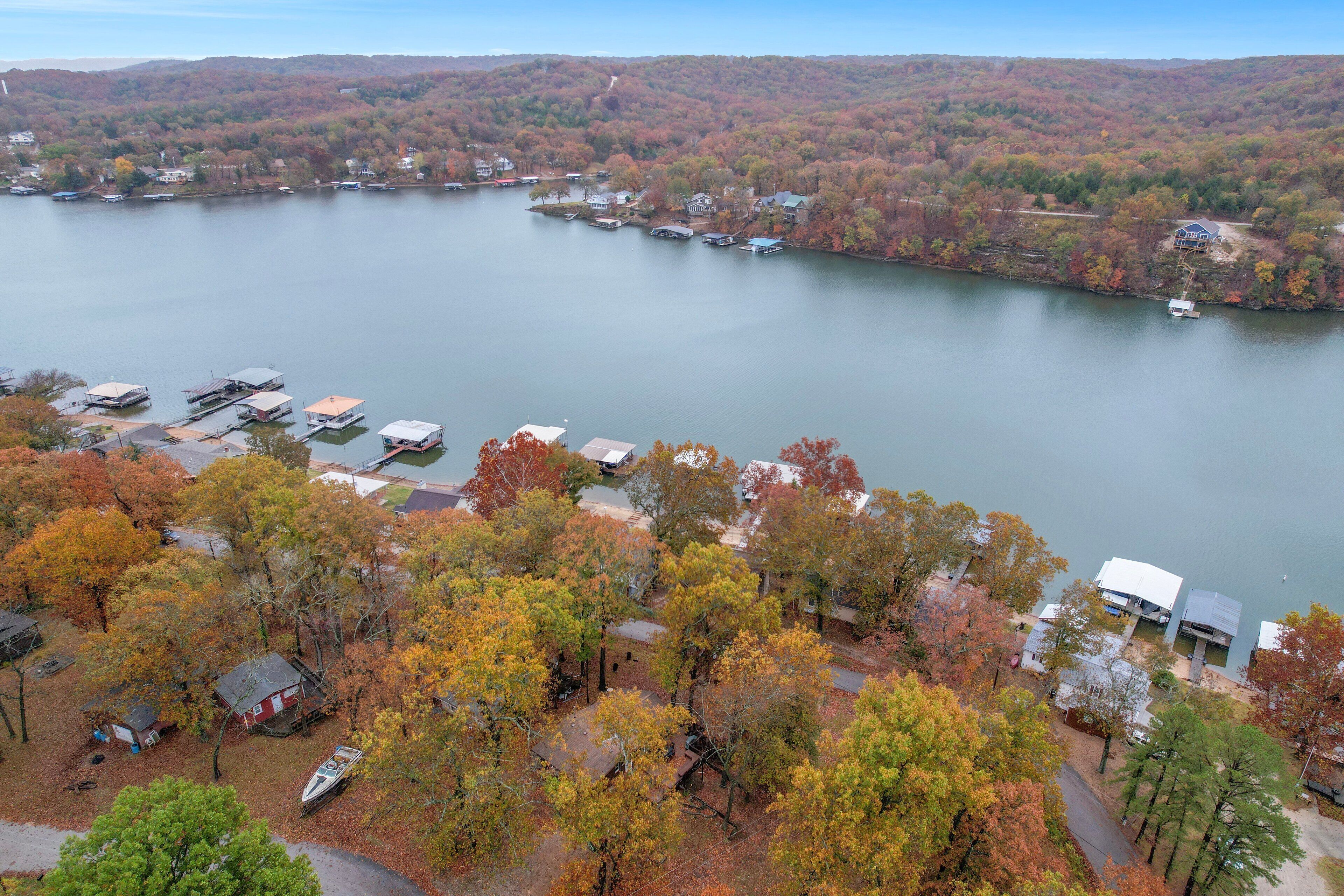 An aerial view of a lakeside property with multiple docked boats, a small marina, and houses surrounded by autumn-colored trees.