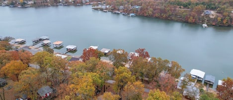 An aerial view of a lakeside property with multiple docked boats, a small marina, and houses surrounded by autumn-colored trees.