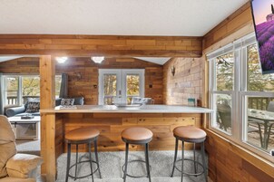 Acogedor interior de cabaña con detalles de madera, una zona de cocina con taburetes y amplias ventanas que ofrecen vistas al exterior.