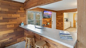 A kitchen with wooden walls, a bar area with stools, a TV mounted on the wall, and a view of the outside through the window. - Stunning Lake View On A Nice & Quiet Relaxing Home (Climax Springs)