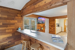 Una cocina con paredes de madera, una barra con taburetes, un televisor montado en la pared y vistas al exterior a través de la ventana.