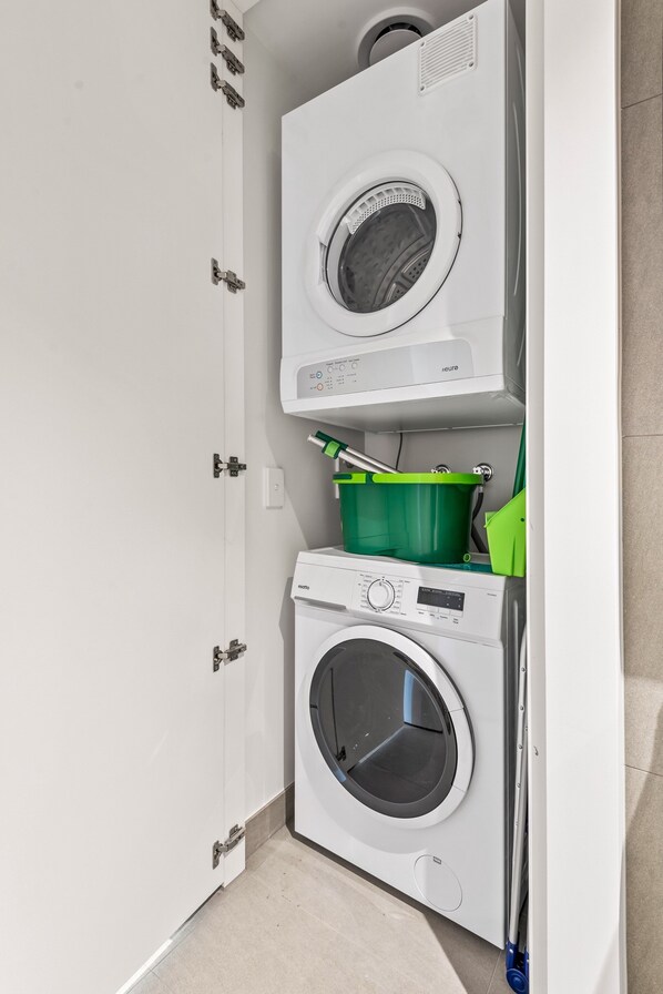 Une petite buanderie comprenant deux machines à laver superposées, un seau-mop vert et une porte blanche.
