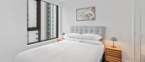 A modern bedroom with a large bed, a nightstand with a lamp, a window with a view of buildings, and a framed abstract painting on the wall.