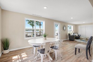 A spacious room with a large dining table, white chairs, and a view of the outdoors through large windows. - Two Homes, Large Deck, Firepit, Central Location (Sunrise Beach)