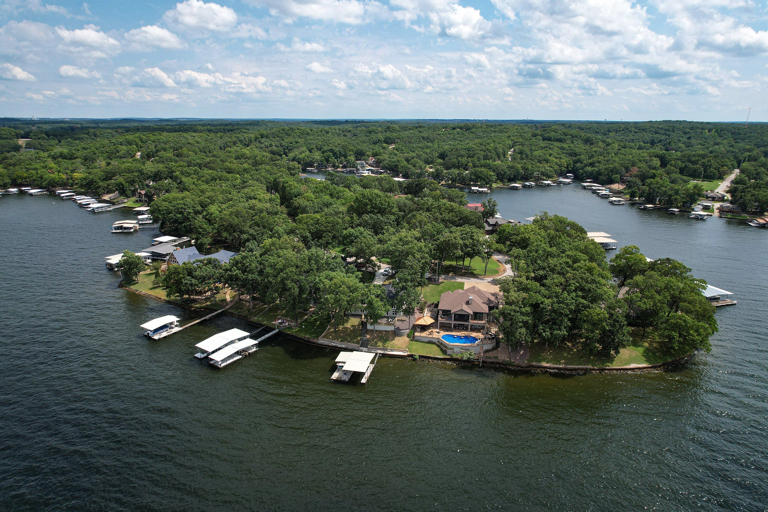 An aerial view of a lakeside property with multiple boats docked, a swimming pool, and lush greenery.