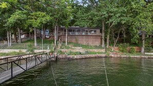 A riverside property with a wooden deck, a covered outdoor area, and a building with a modern design. - Quiet Cove, Private Dock, Fishing, Swimming, Boat Slip & Kayaks (Camdenton)