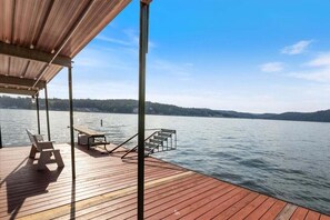 A wooden deck with a covered area, a chair, a bench, and a staircase leading to a dock.