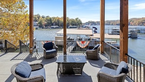 Un espace extérieur aménagé, abrité et doté de mobilier en osier et d’une table, offrant une vue sur un lac où l’on peut apercevoir des bateaux et des embarcations amarrées.