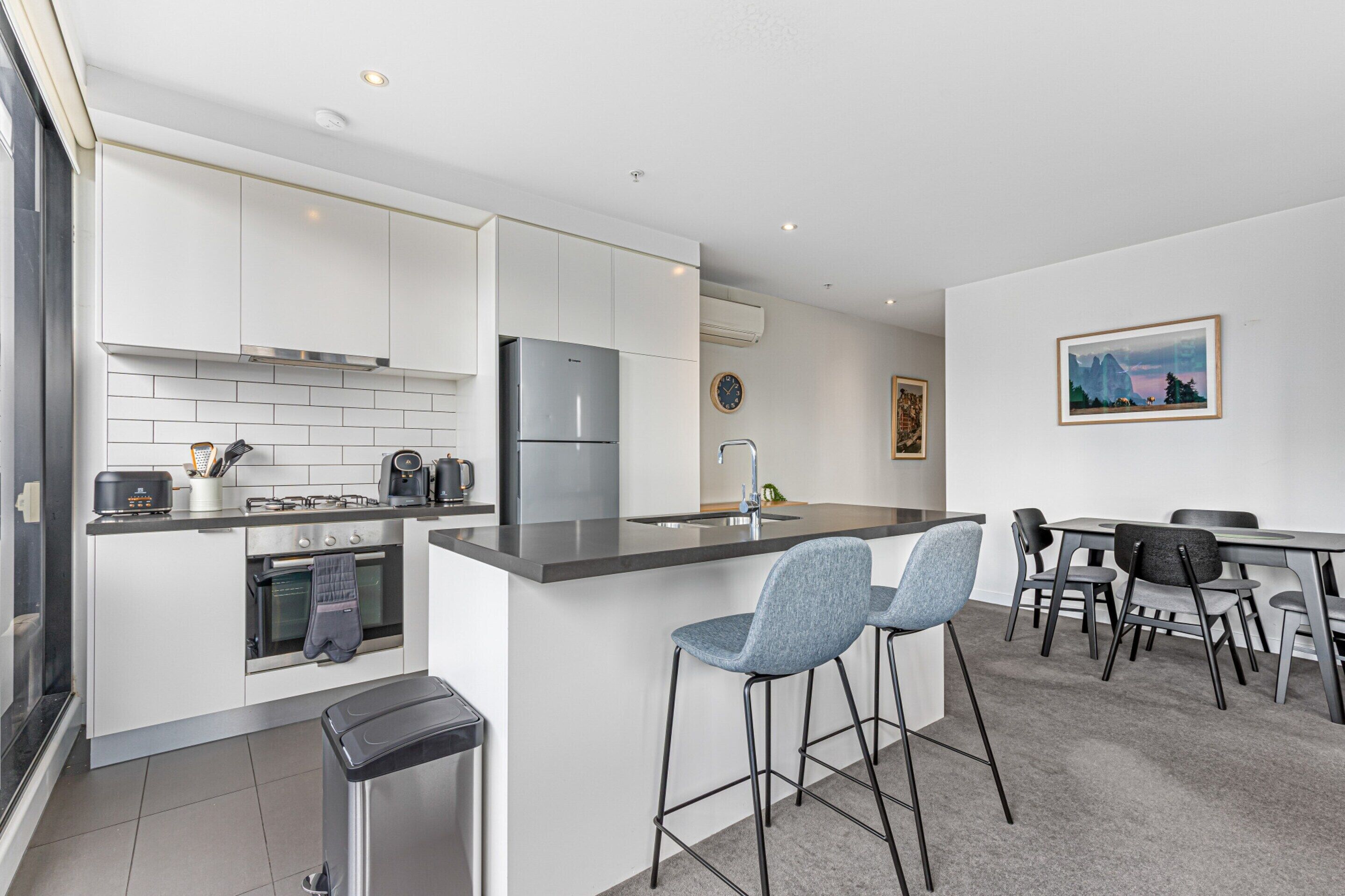 A modern kitchen with white cabinets, a stainless steel refrigerator, and a built-in oven. There are two bar stools and a dining table with chairs.