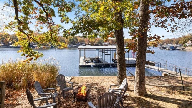 A lakeside deck with Adirondack chairs, a fire pit, and a dock extending into the water.