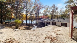 A house with a sandy yard, trees with autumn foliage, and a body of water in the background.