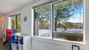 A room with a large window overlooking a body of water and trees, a white brick wall, and a white shelf unit with folded clothes.