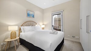 A bedroom with a bed, bedside lamps, a wicker side table, a painting on the wall, and a door with a view. - Spacious 3-bed Townhouse Near Dromana Beach (Melbourne)