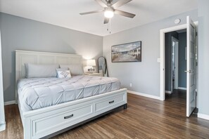 Un dormitorio con cama, mesitas de noche, ventilador de techo y un pasillo que conduce a otra habitación.
