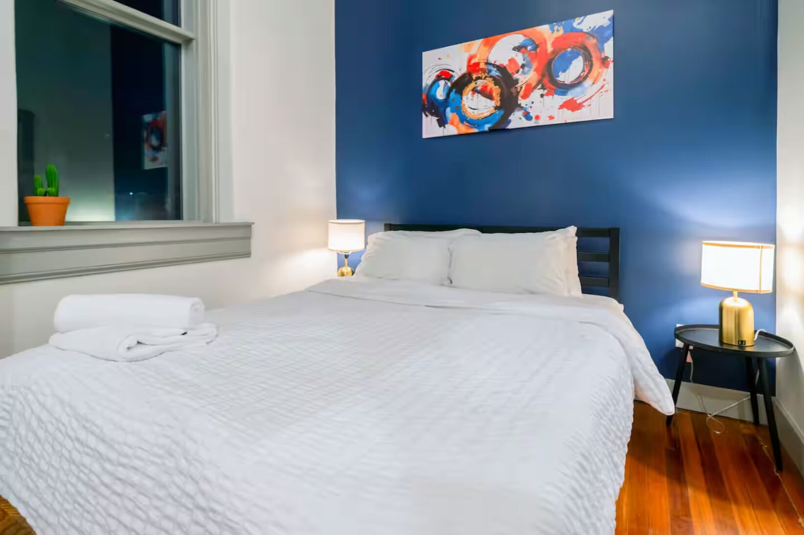 Een net opgemaakt bed met witte lakens, een blauwe accentmuur en een kleurrijk abstract schilderij erboven.