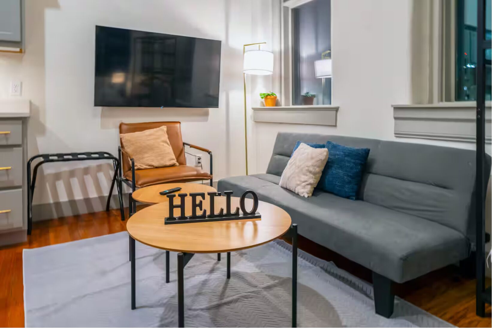 A modern living room with a grey sofa, a wooden coffee table with 'HELLO' letters, a brown leather chair, a TV mounted on the wall, and a window with a view of the city.