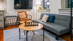 Een moderne woonkamer met een grijze bank, een houten salontafel met de tekst 'HELLO', een bruine lederen fauteuil, een aan de muur bevestigde televisie en een raam met uitzicht op de stad.