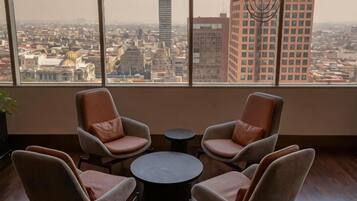 Quarto com quatro poltronas, uma mesa pequena, amplas janelas e vista para a cidade, além de um lustre.