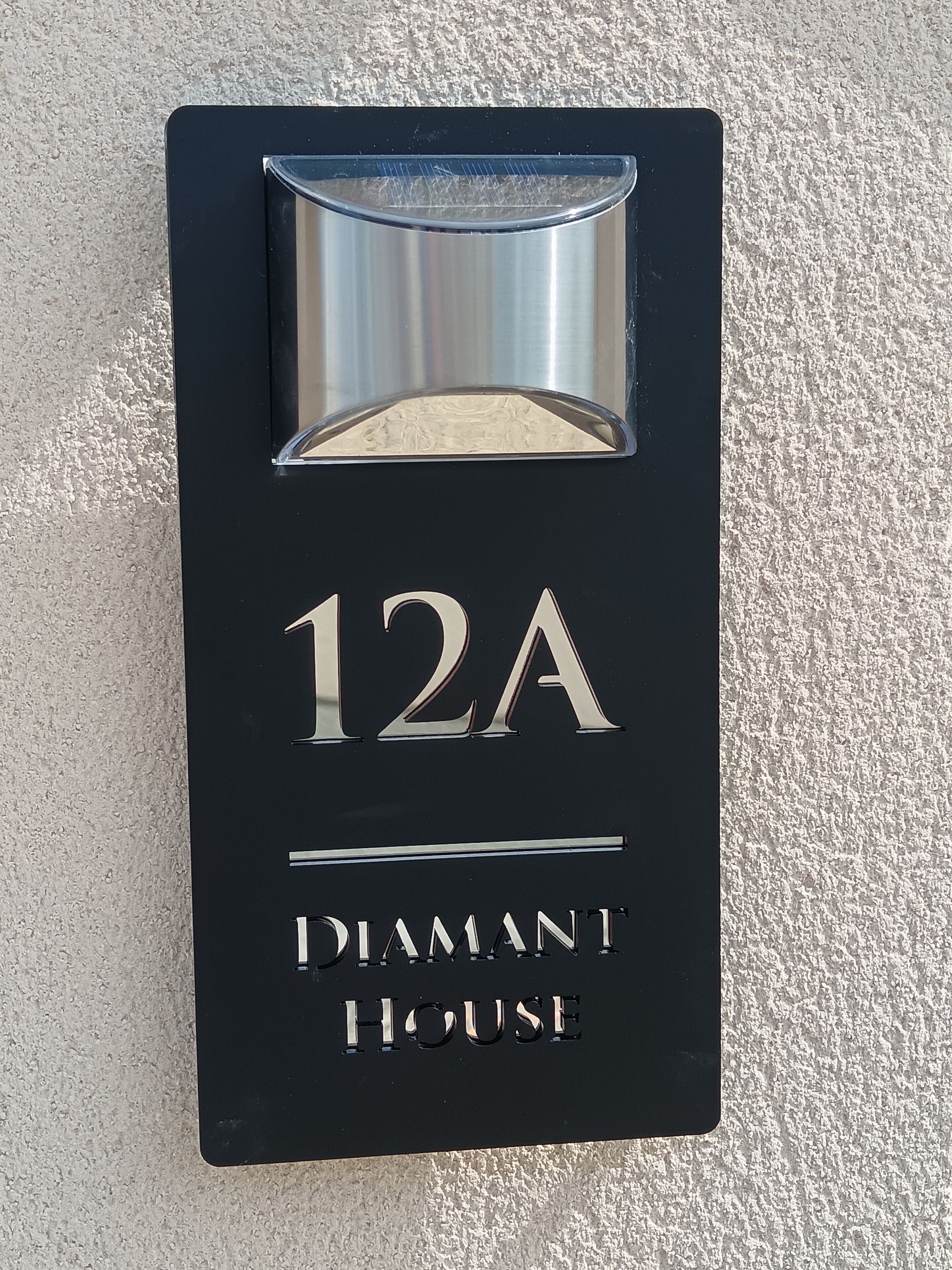 Une plaque noire portant le numéro de maison « 12A » et l’inscription « DIAMANT HOUSE », le tout gravé sur la plaque.