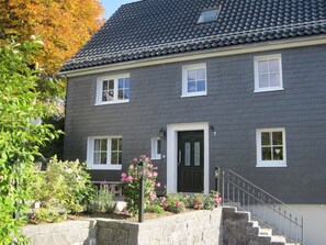 Une maison à deux étages, avec une façade de couleur gris foncé, des fenêtres blanches, une porte d’entrée noire et un escalier en pierre menant à l’entrée.