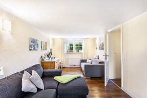 Un salon avec un canapé gris, un plus petit canapé gris, une table d’appoint en bois et une fenêtre avec des rideaux.