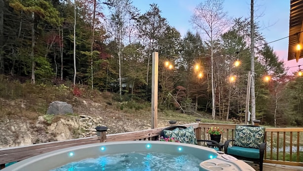 Un bain à remous doté d’une terrasse en bois et illuminé de guirlandes lumineuses, entouré d’arbres et bordé d’une colline rocheuse.