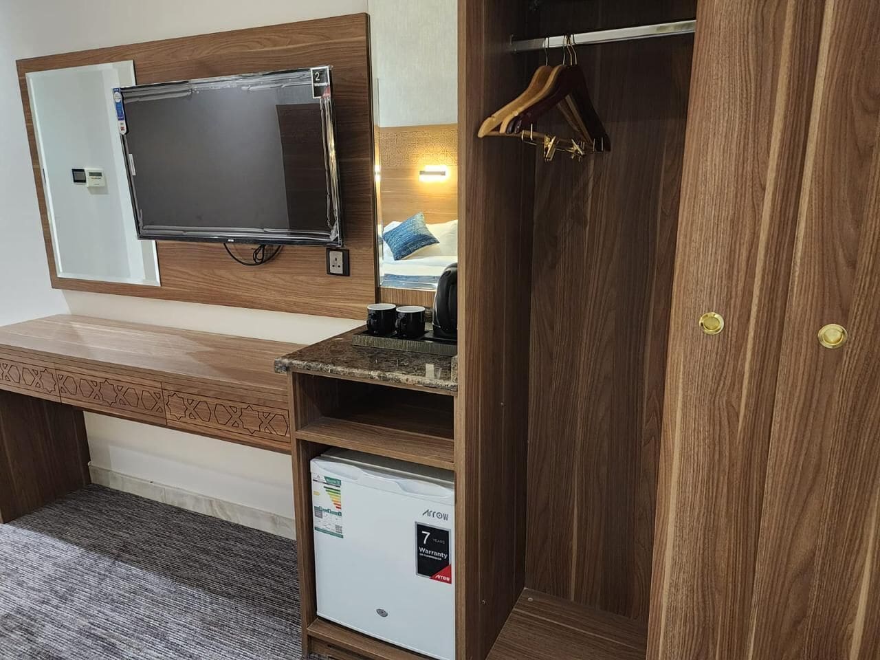 A compact hotel room with a wooden desk, a television, a wardrobe, and a mini-fridge.