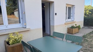 Une terrasse avec une table et des chaises vertes, des jardinières en bois ornées de plantes, et un bâtiment blanc doté de fenêtres et d’une porte.
