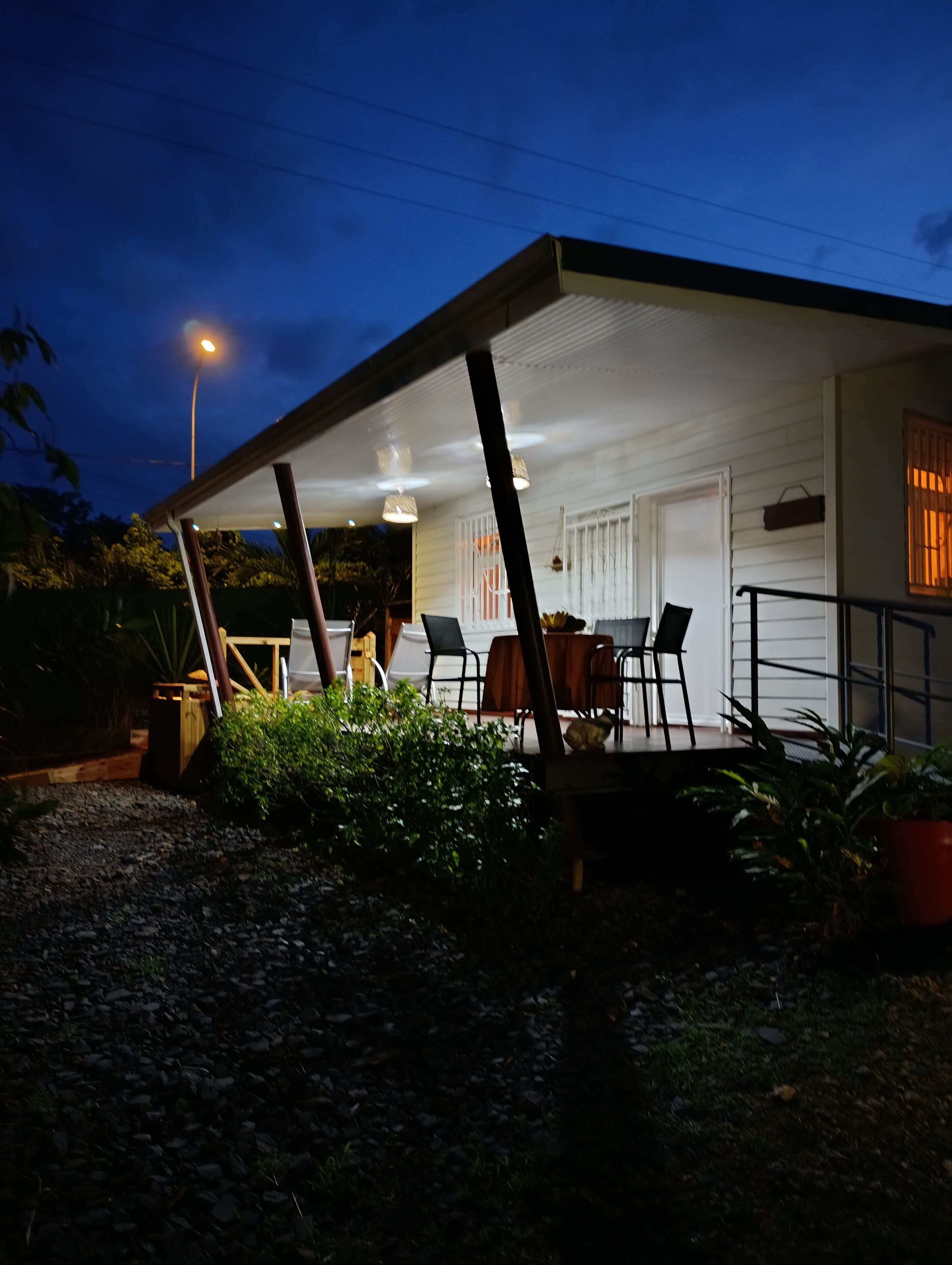 A covered outdoor area with a table and chairs, surrounded by plants and a gravel path.