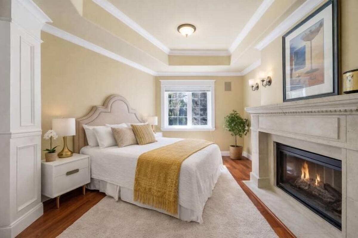 A bedroom with a bed, nightstand, lamp, fireplace, window, and a framed picture on the wall.