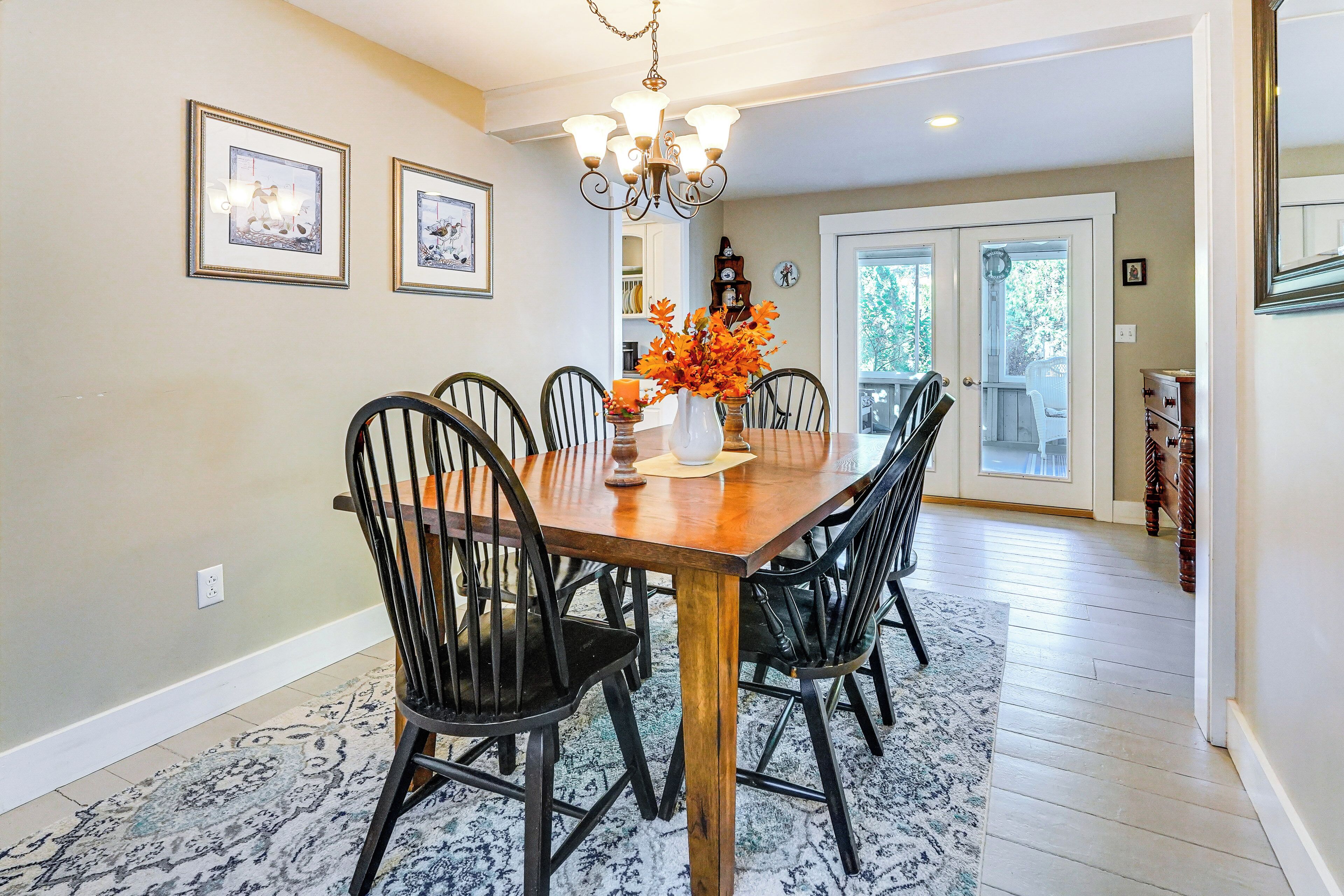 A dining area with a wooden table, black chairs, a chandelier, framed artwork on the wall, and a patterned rug.