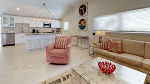 Una sala de estar moderna con un sofá, un sillón rojo estampado, una mesa de centro y una zona de cocina con armarios blancos y un microondas.