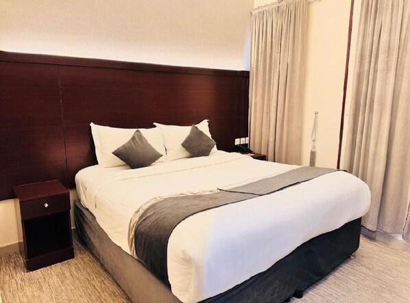 Una habitación de hotel con una cama grande, cabecera de madera oscura, ropa de cama blanca y una mesita de noche pequeña.