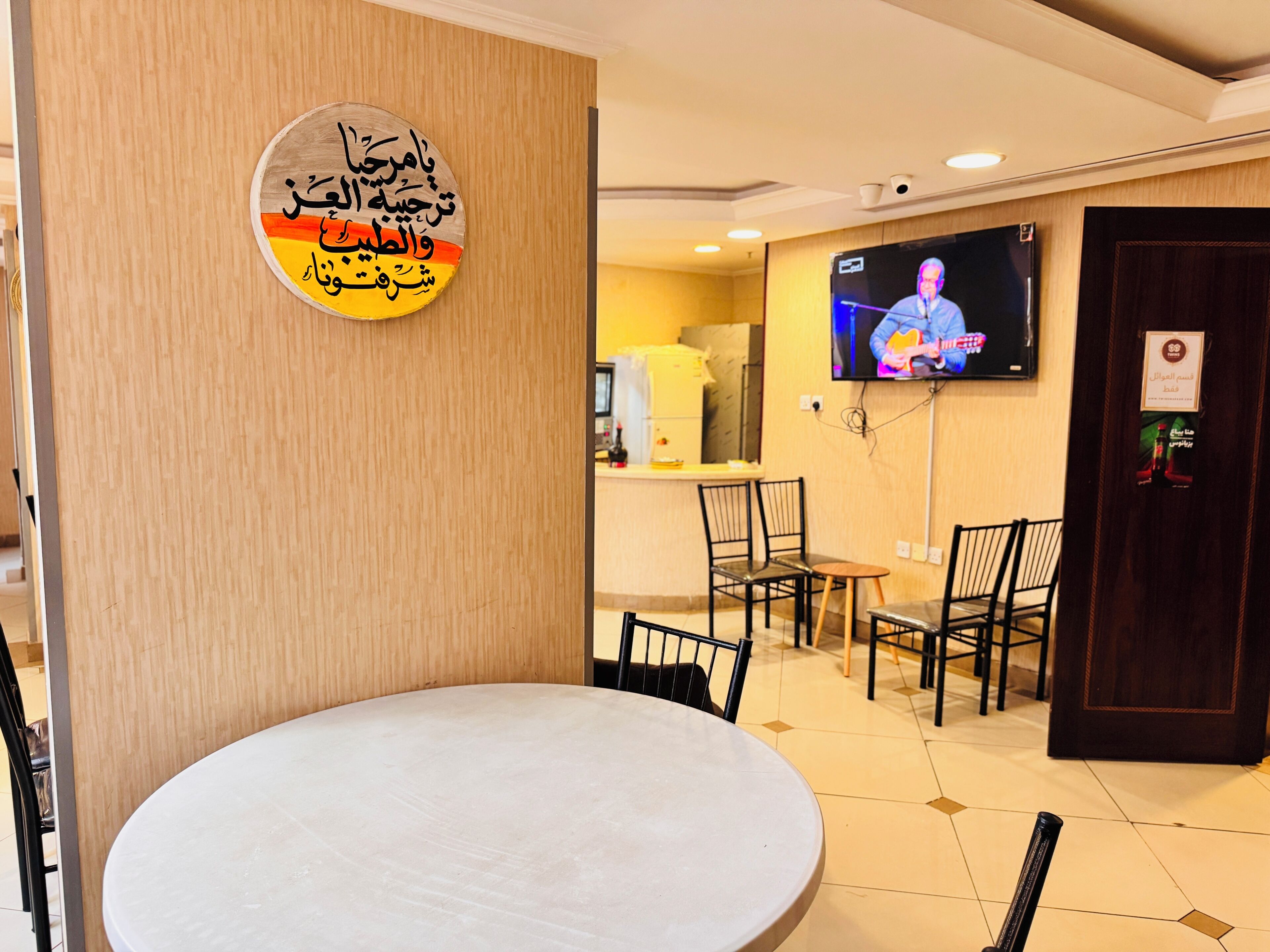 A hotel lobby with a round table, chairs, a television mounted on the wall, and a wooden panel with Arabic calligraphy.