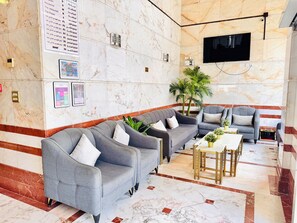 A modern lobby with marble flooring, grey sofas, a flat-screen TV, and a large potted plant. - Rowaa Makarem (Makkah)